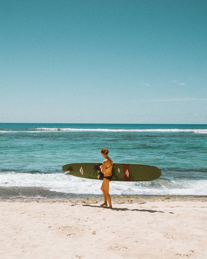 Surfer carries board along sunny Hawaiian beach with clear blue ocean.