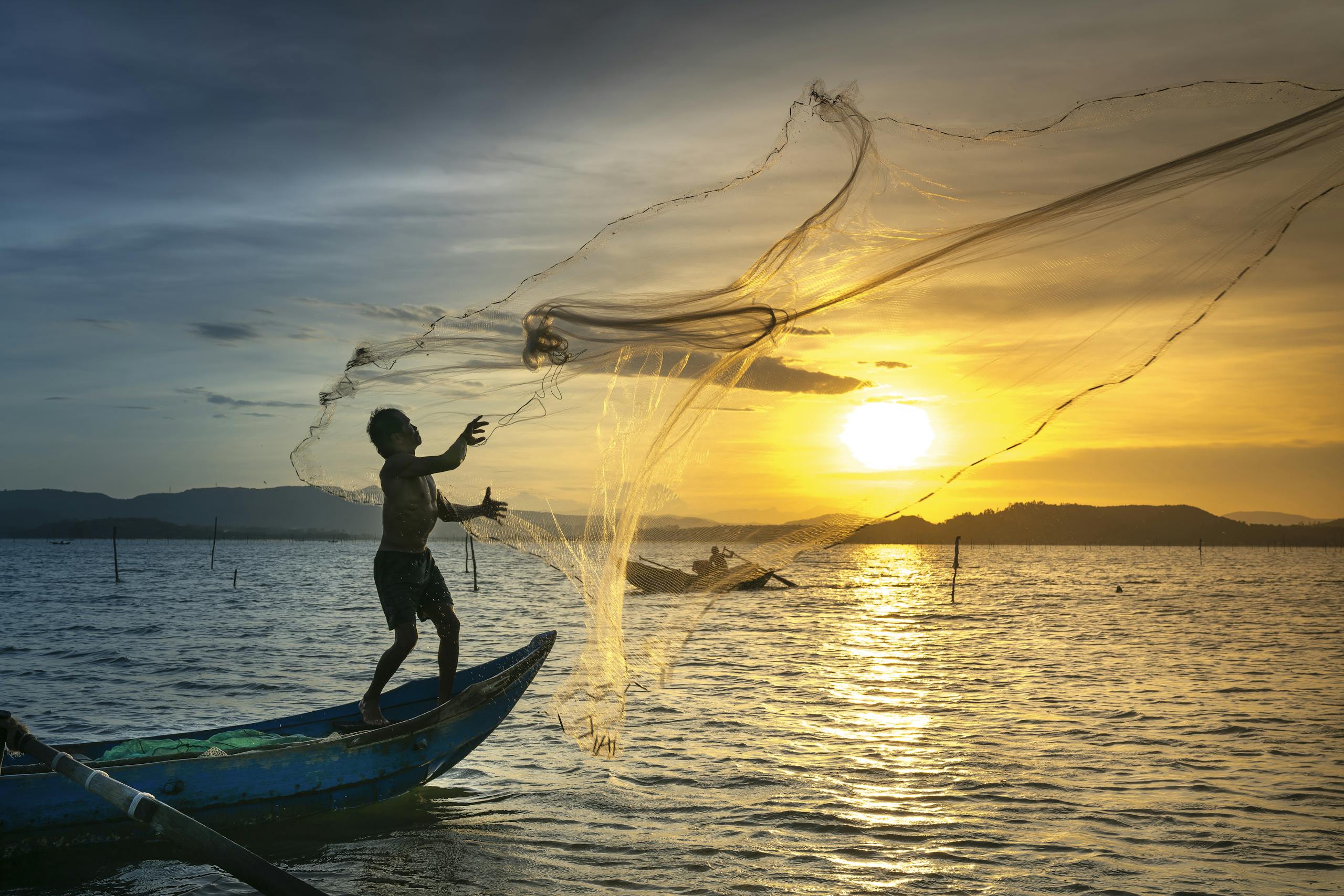 A fisherman skillfully casts a net at sunrise, silhouetted against a golden sky over a calm lake.