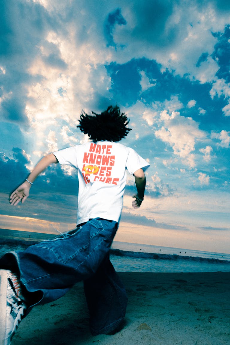 A man with curly hair runs on the beach under a dramatic cloudy sky, wearing a graphic T-shirt.