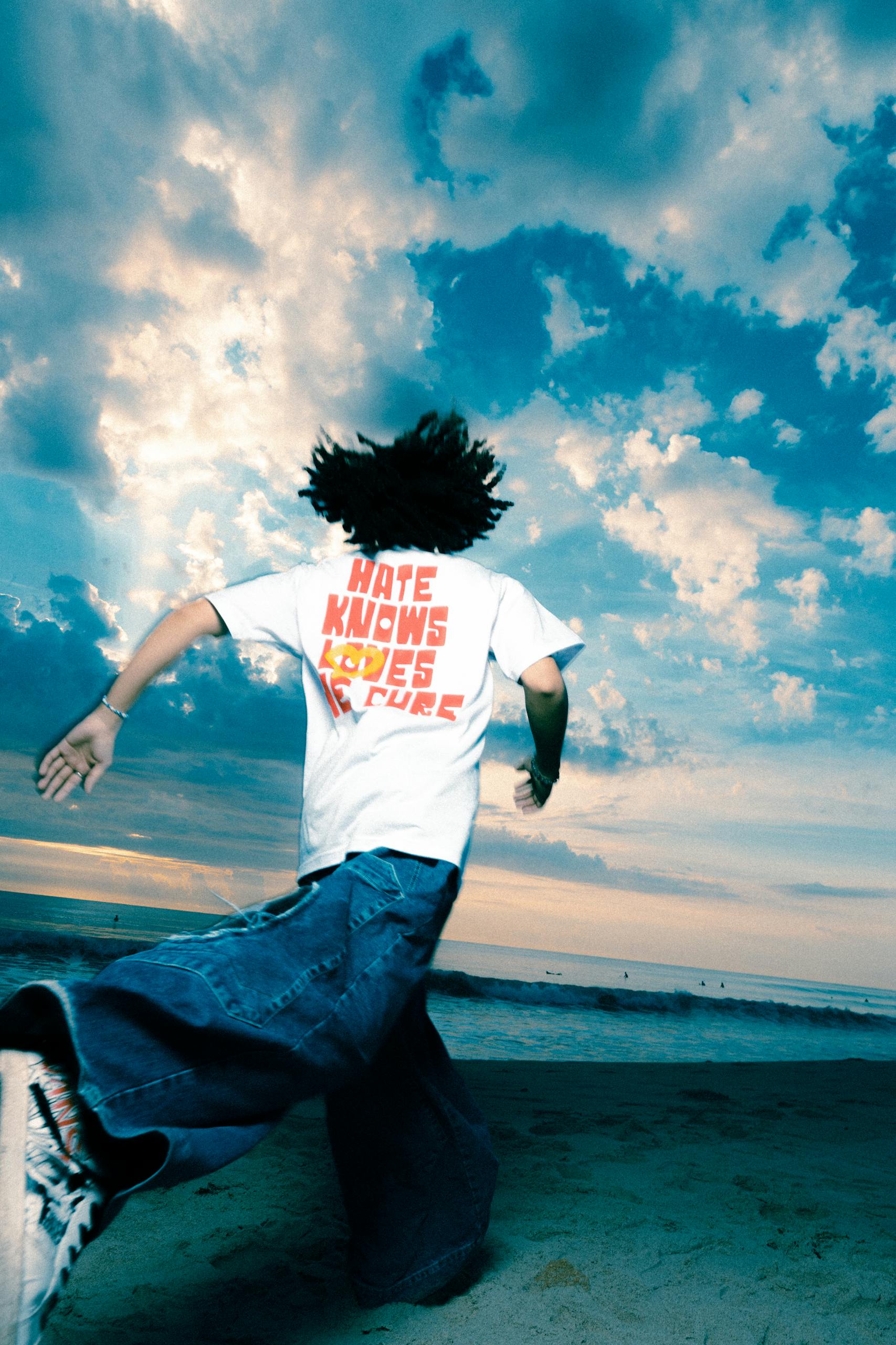 A man with curly hair runs on the beach under a dramatic cloudy sky, wearing a graphic T-shirt.