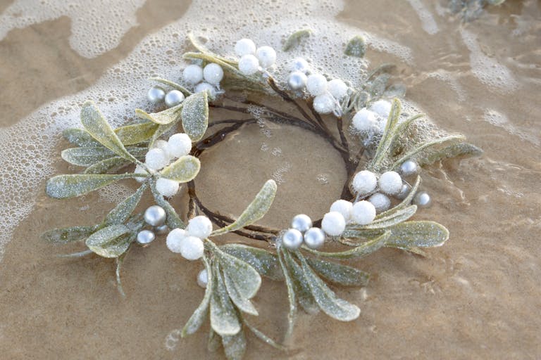A Christmas wreath with ornaments on a sandy beach, touched by gentle waves.